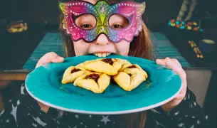 ילדה עם מסיכה מחזיקה צלחת עם אוזני המן