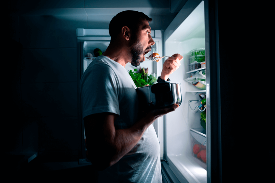 An adult takes a pot out of the fridge and eats with a spoon in the middle of the night